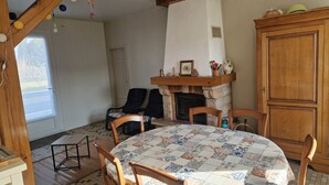Ett mysigt rum med en öppen spis, trästolar, ett runt bord med en mönstrad duk och utsikt över naturen genom ett fönster.