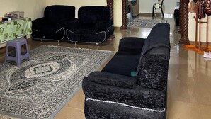 Un salon avec un canapé noir, un tapis à motifs et un petit tabouret. Deux tableaux encadrés ornent le mur et le plafond est en bois.