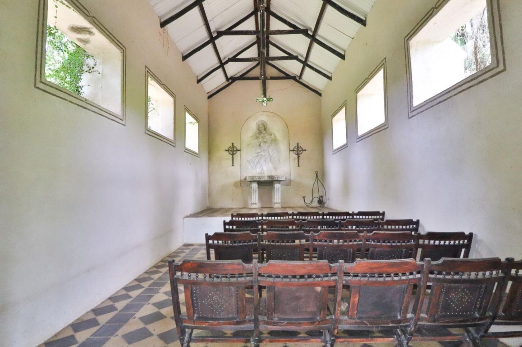 L’intérieur d’une église avec des bancs en bois, un plafond élevé doté d’une structure en poutres apparentes, et un autel central surmonté d’une statue.