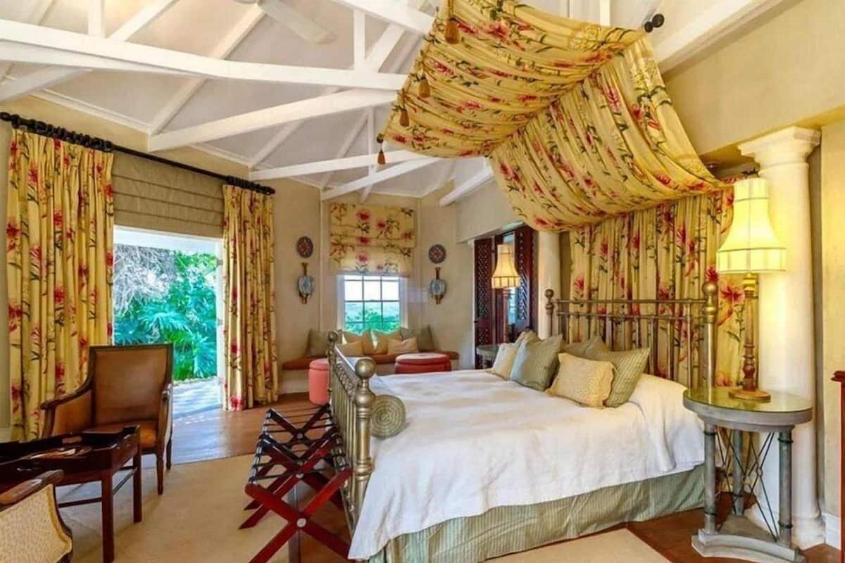 A spacious bedroom with a large bed, a wooden chair, a small table, and a window with a view of greenery.