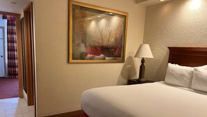 Una habitación de hotel con una cama grande, una mesita de noche con lámpara, un cuadro en la pared y una puerta que da a otra habitación.
