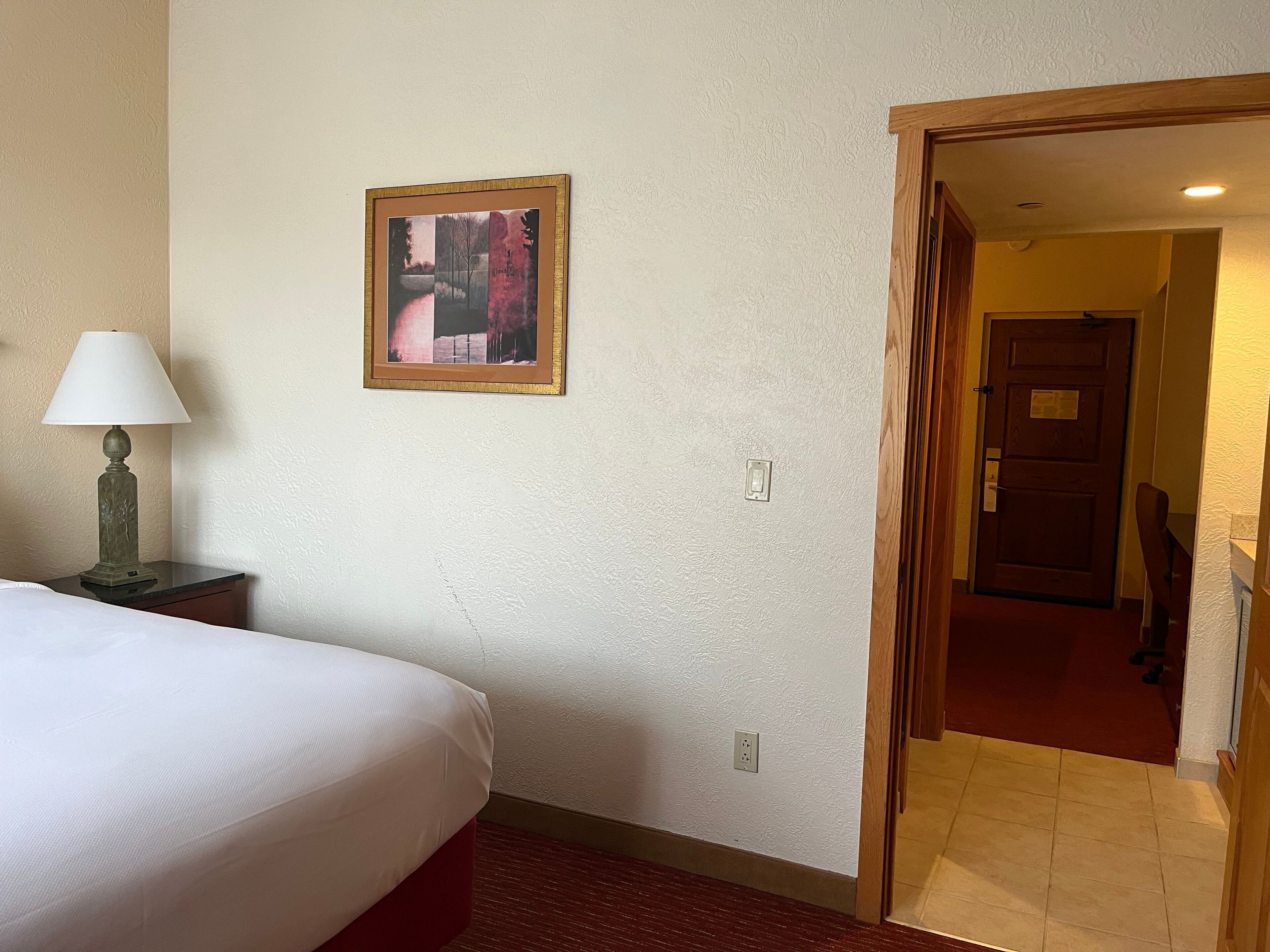 A hotel room with a bed, a nightstand with a lamp, a painting on the wall, and a door leading to another room.