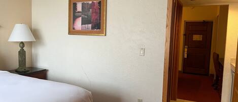 A hotel room with a bed, a nightstand with a lamp, a painting on the wall, and a door leading to another room.
