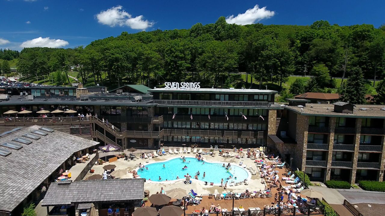 A hotel with a large outdoor pool surrounded by lounge chairs and umbrellas, a covered deck area, and a view of a forested hill.