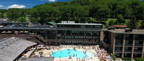 A hotel with a large outdoor pool surrounded by lounge chairs and umbrellas, a covered deck area, and a view of a forested hill.