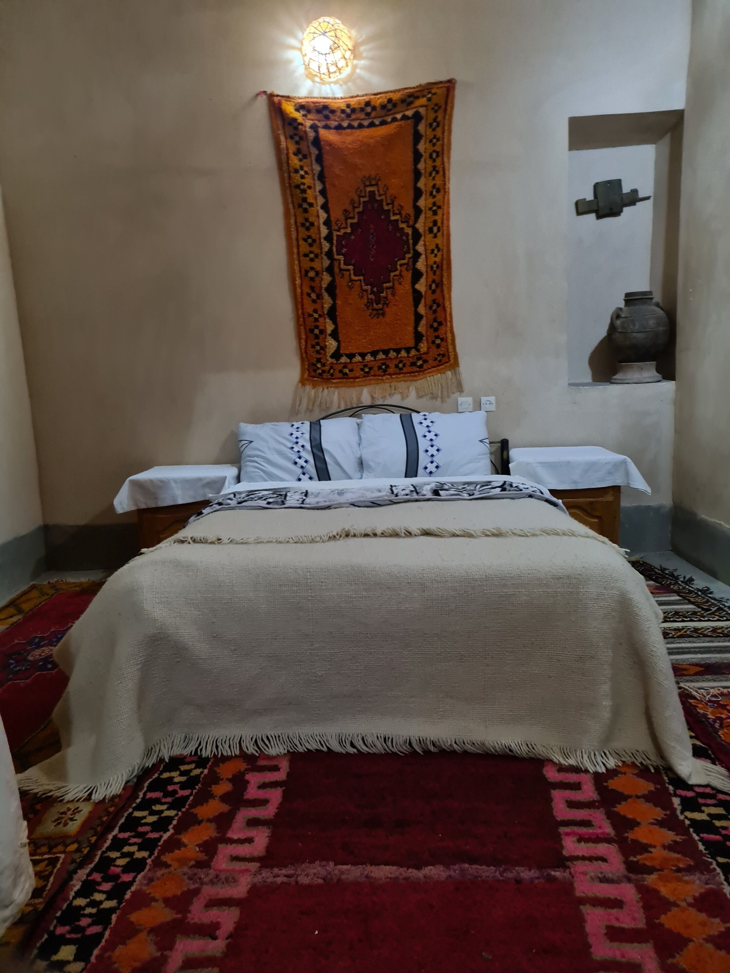 Ein Bett mit weißer Bettdecke und Kissen, ein Teppich mit roten und orangefarbenen Mustern sowie eine wandmontierte Lampe.