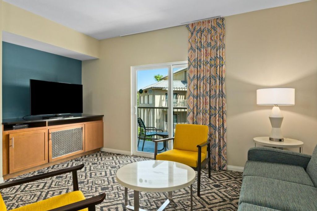 A hotel room with a television, a sofa, a yellow armchair, a small table, and a balcony with a patterned curtain.