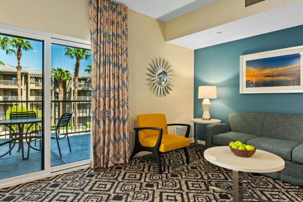 A hotel room with a balcony, a patterned carpet, a sofa, a chair, a table with a fruit bowl, and a wall-mounted lamp.