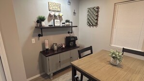 Un comedor con una mesa y sillas de madera, una estación de café con tazas y cafetera, y un estante de pared con objetos decorativos.