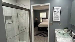 Un baño moderno con cabina de ducha de vidrio, tocador con azulejos de mármol y un cartel con las normas del baño enmarcado.