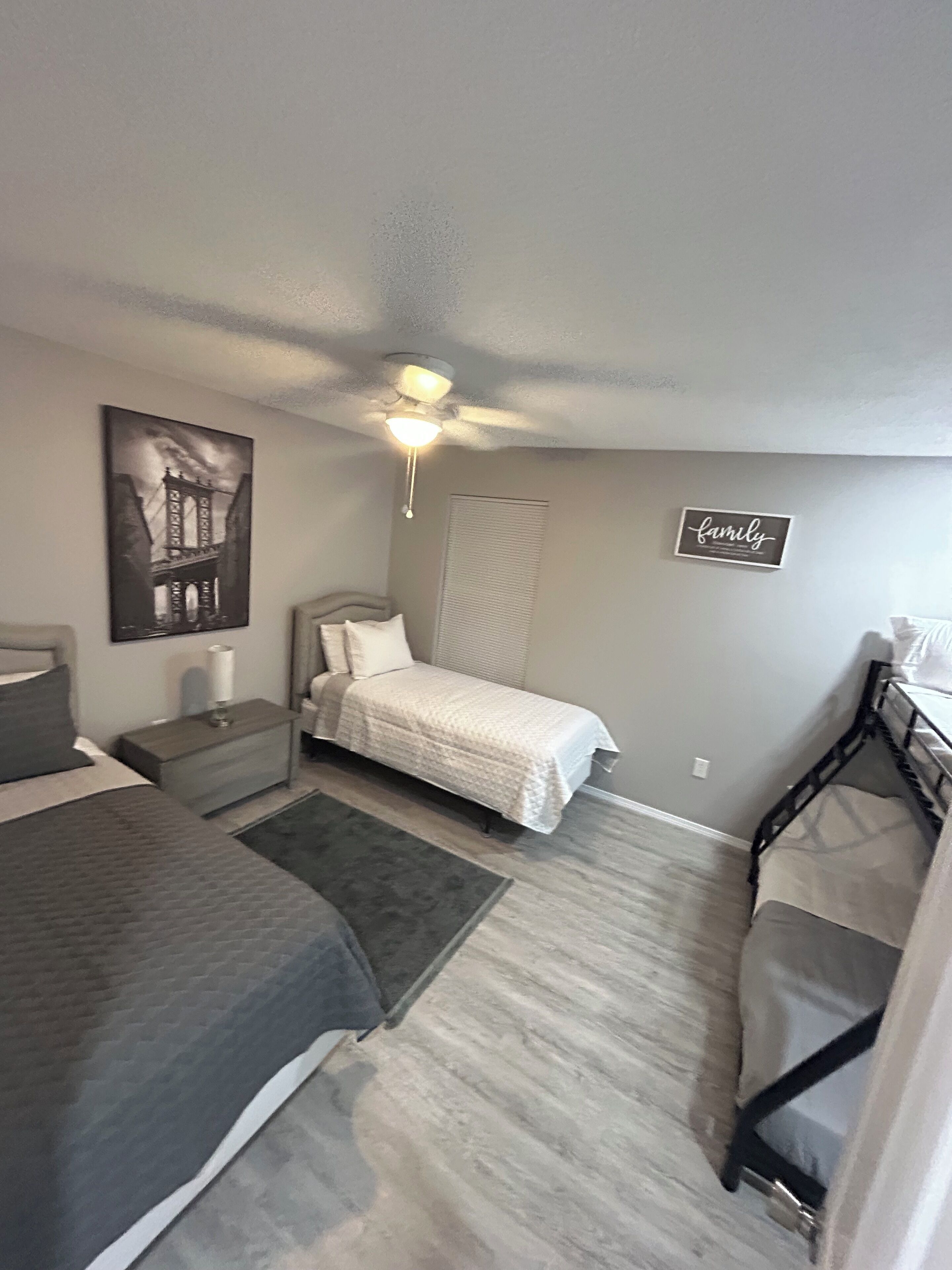 A bedroom with a bed, a nightstand, a bedside table, a wall-mounted picture, a ceiling fan, and a 'family' sign.