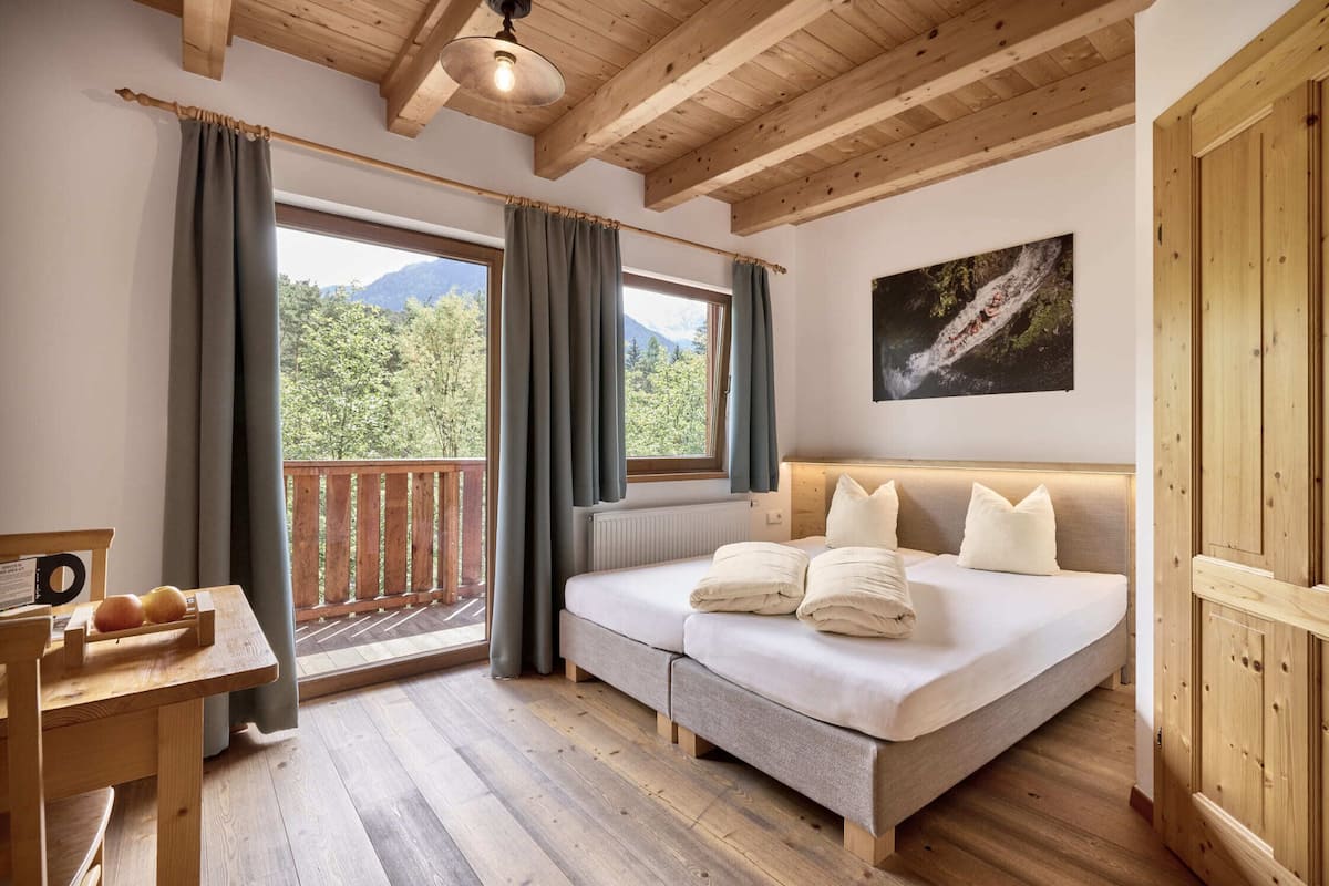 Ein Schlafzimmer mit Holzbalkendecke, einem Bett mit Kissen, einem Tisch mit Obst und Blick auf Berge durch das Fenster.