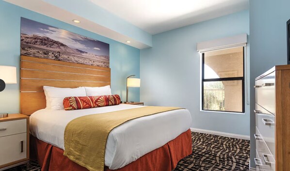 A hotel room with a large bed, a nightstand, a lamp, a window with blinds, and a view of a landscape.