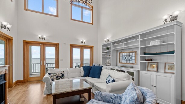 Amplia sala de estar con techo alto, ventanas grandes y vista al mar. Hay dos sofás blancos con cojines azules, un sillón azul con estampado y una mesa de centro de madera. Contra la pared, un mueble blanco con estantes.