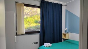 Une chambre à coucher avec un lit, une fenêtre avec des rideaux, une table de chevet avec une lampe, et une vue sur la verdure à l’extérieur.
