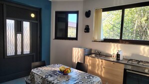 Une cuisine compacte avec un coin repas, comprenant une table agrémentée d’un panier de fruits, une fenêtre avec des stores et une vue sur les arbres à l’extérieur.