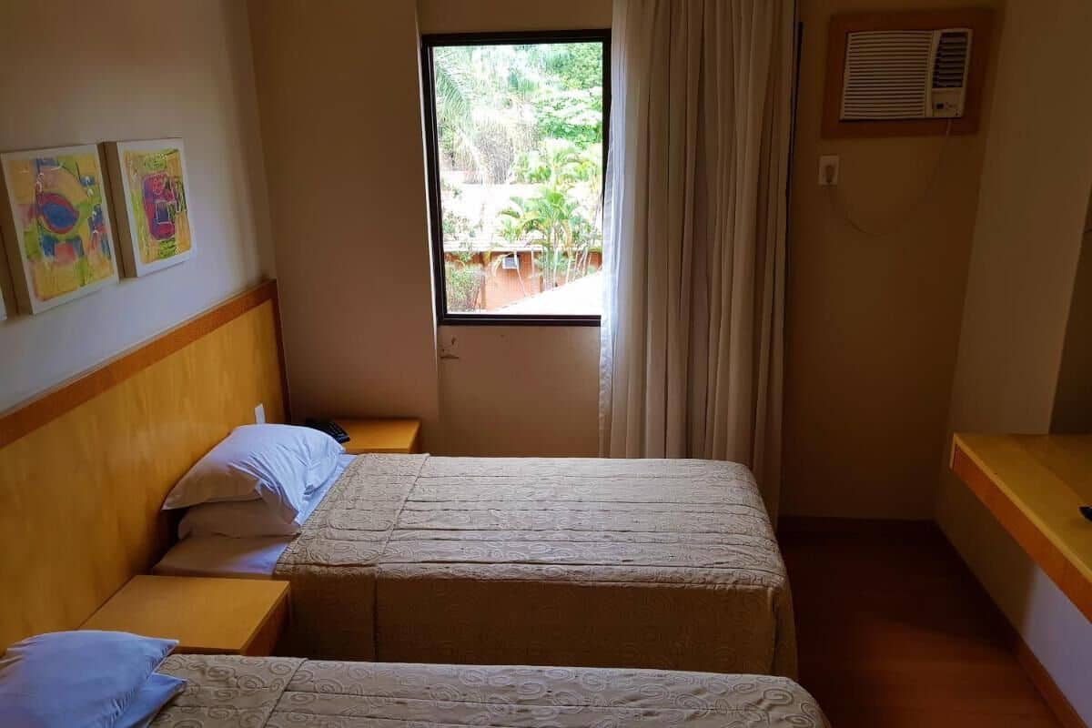 A hotel room with two beds, a window with curtains, a wooden headboard, and a small table with a lamp.