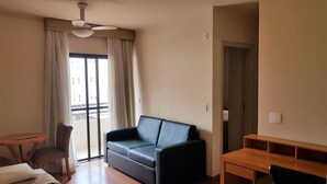 A hotel room with a bed, a black leather sofa, a wooden desk, and a chair. There is a window with curtains and a door to the right. - Ômega Flat (Bauru)