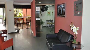 A reception area with a black leather sofa, a small table with a white orchid, and a glass partition separating the reception from the dining area. - Ômega Flat (Bauru)