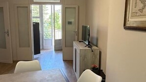Un salon moderne comprenant une table à manger, deux chaises, un téléviseur posé sur un meuble en marbre et une grande fenêtre donnant sur un paysage verdoyant.