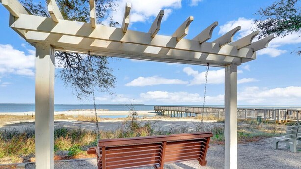 Eine hölzerne Schaukelbank unter einer Pergola mit Blick auf den Strand und einen Holzsteg.