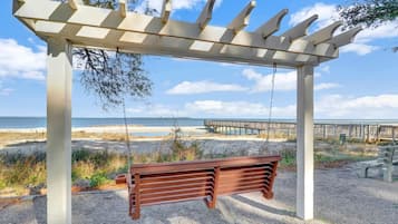 A wooden swing bench under a pergola with a view of the beach and a wooden pier.