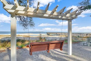 A wooden swing bench under a pergola with a view of the beach and a wooden pier.