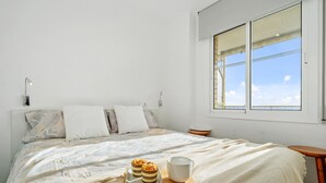 Un dormitorio con una cama, almohadas, una mesita de noche, una ventana y una bandeja con un pastel y una taza.