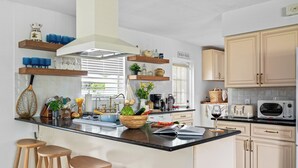 A modern kitchen with a central island, bar stools, wooden shelves, and a sign reading "BEACH HOUSE".