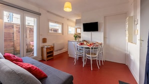 A compact living space with a dining area, a sofa with red cushions, a television mounted on the wall, and a sliding glass door leading to an outdoor area.