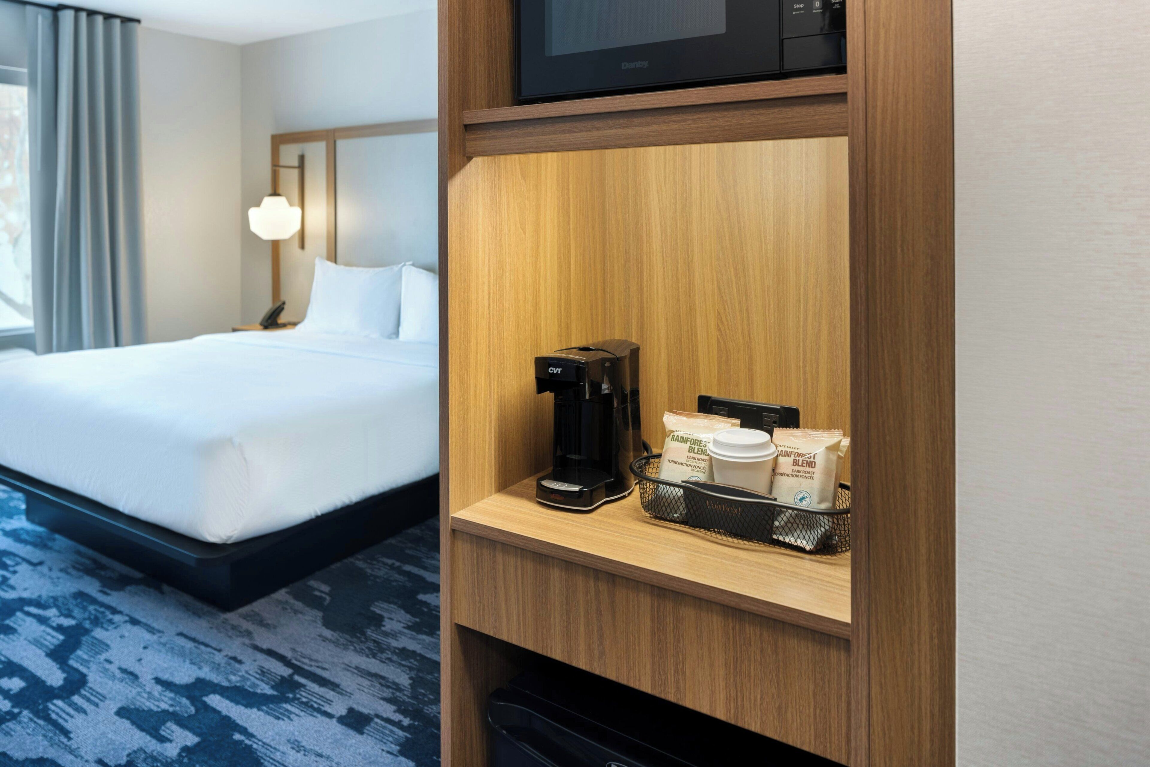 A hotel room with a bed, a coffee maker, a microwave, and a shelf with coffee cups and a bag of coffee.