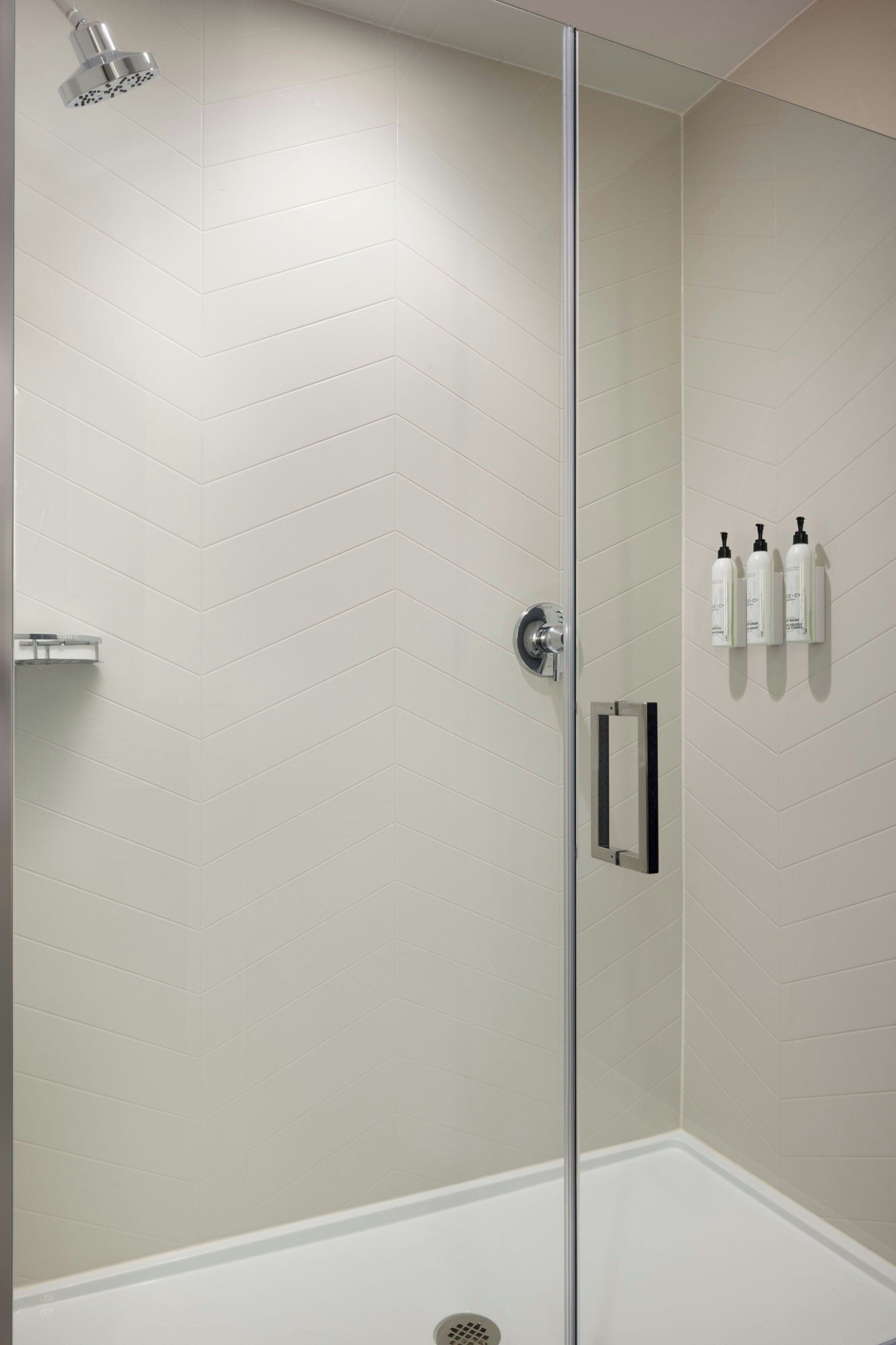 A modern shower with a glass door, a white tiled wall, and a small shelf with toiletries.