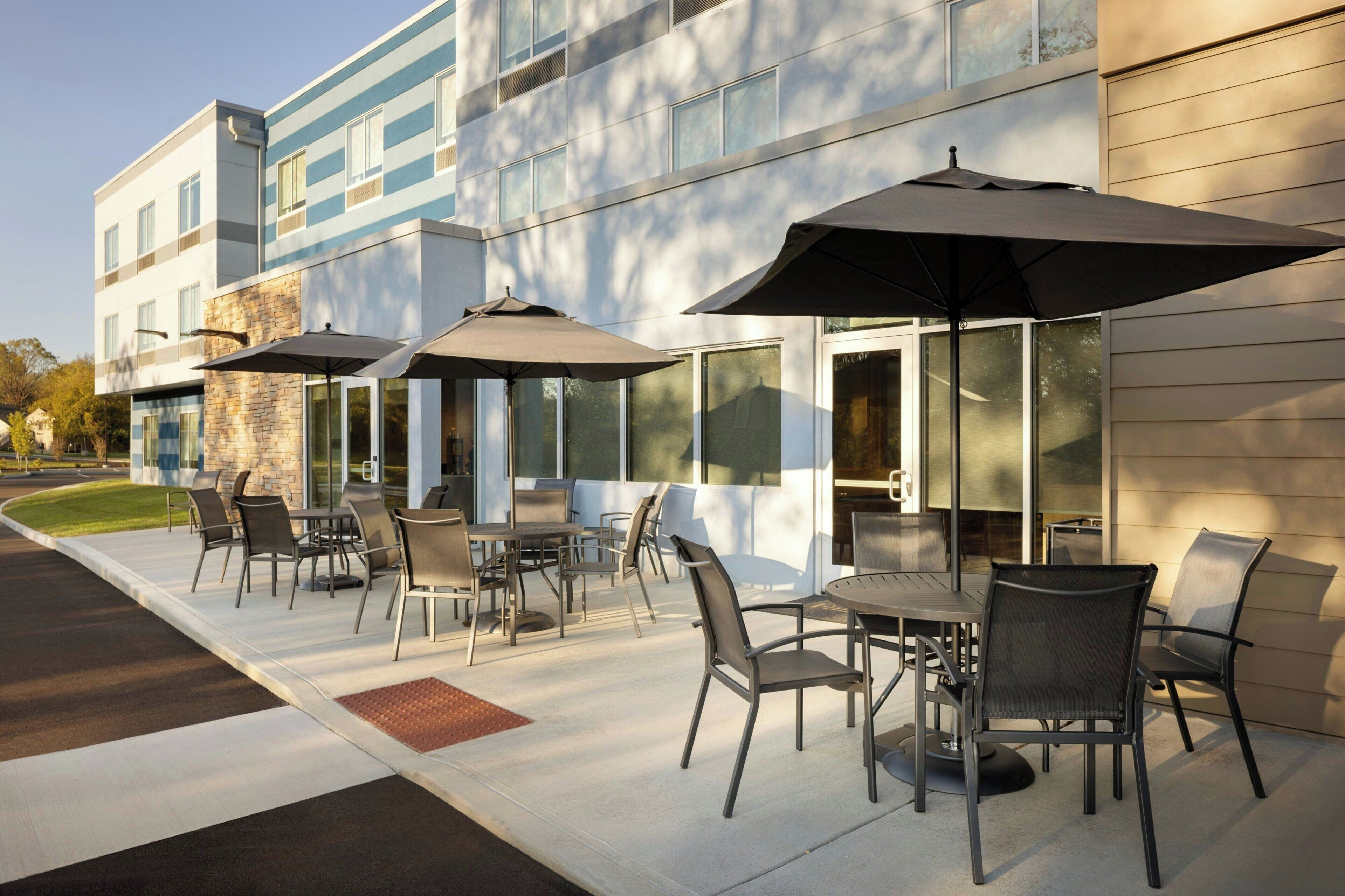 Une terrasse extérieure avec des tables et des chaises, de grands parasols, et un bâtiment doté de grandes fenêtres et d’une façade en pierre.