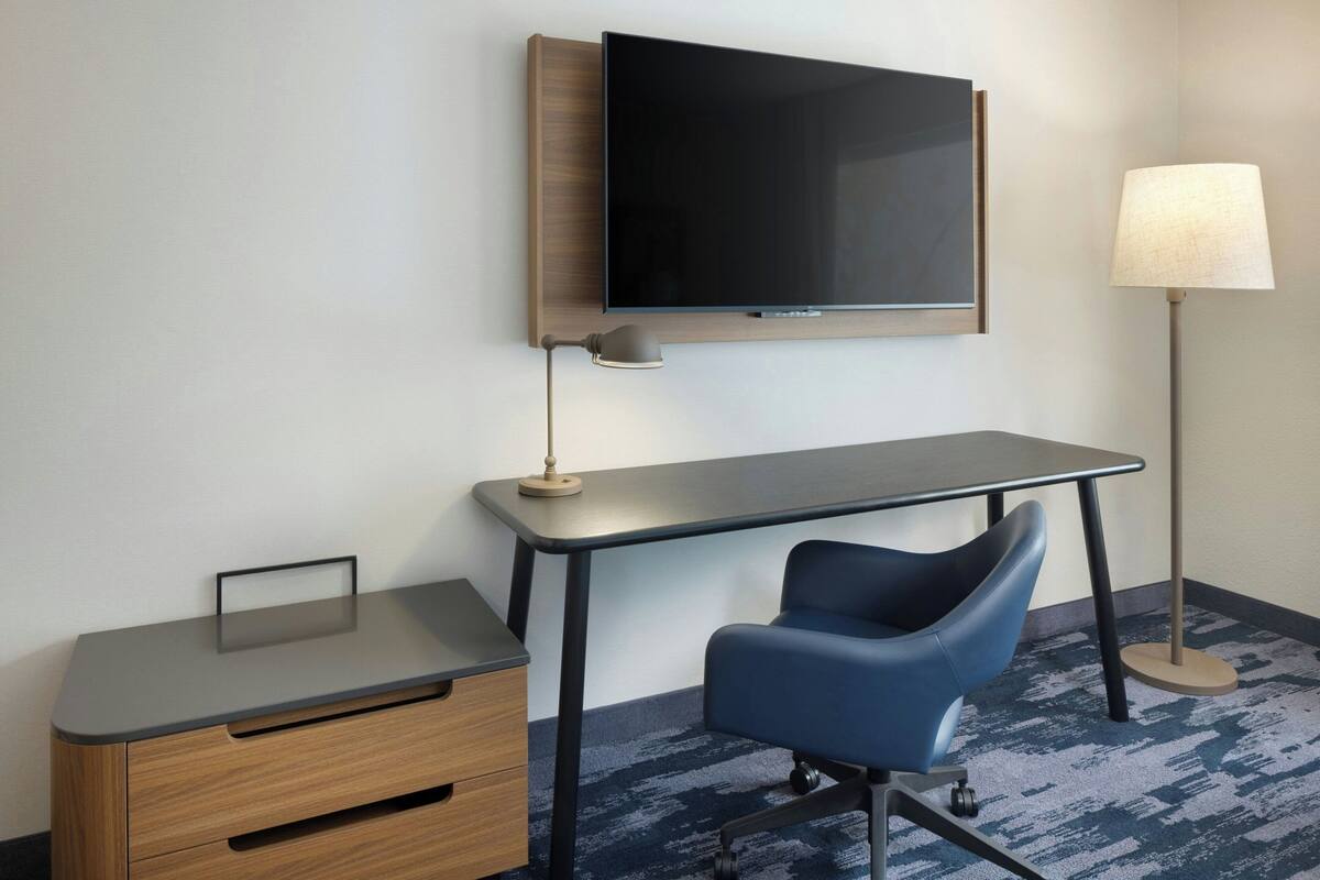 Habitación de hotel con escritorio, silla, televisor, lámpara y una mesita.