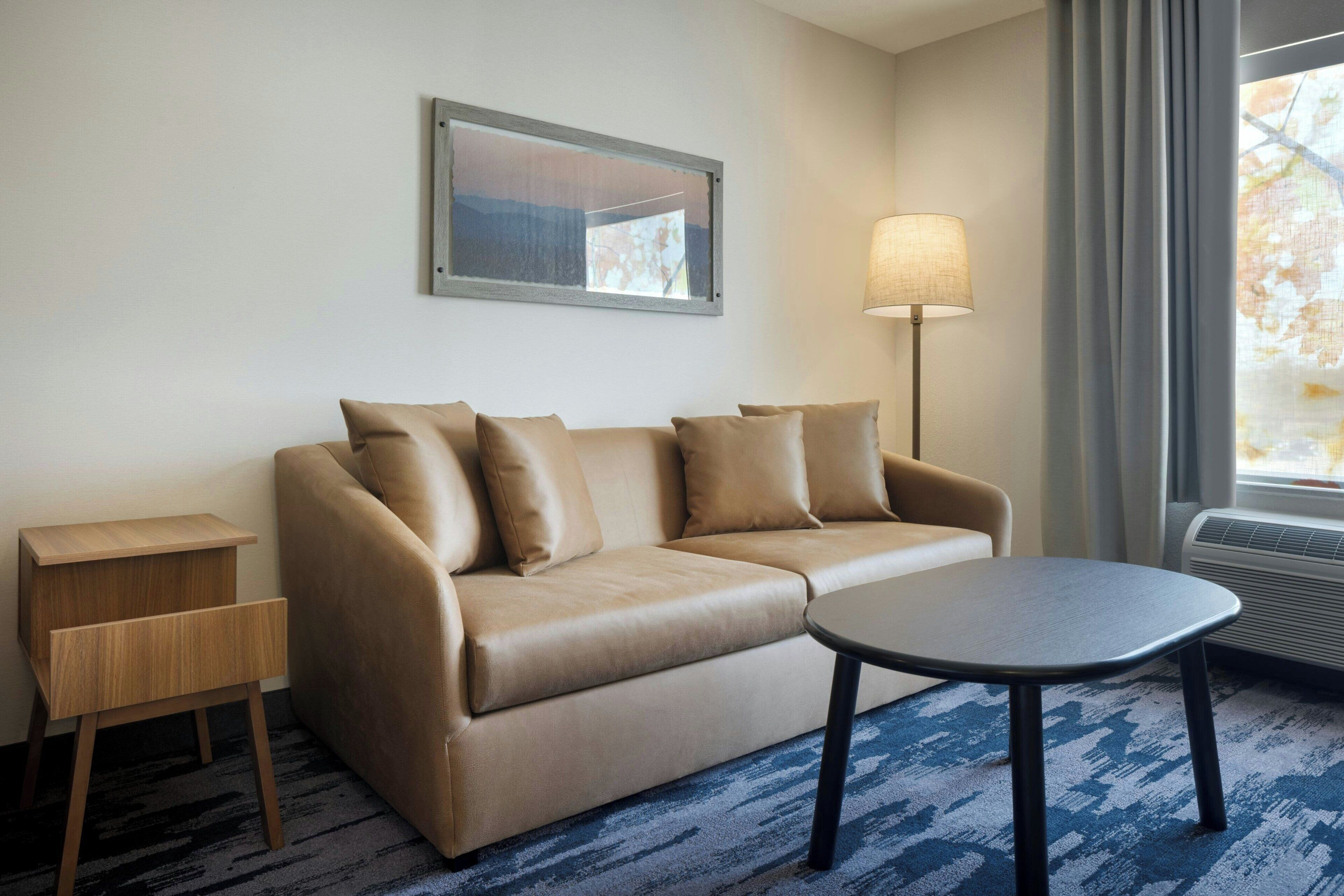 Habitación de hotel con un sofá color beige, una pequeña mesita de madera, una mesa redonda de centro, una lámpara de pie y un amplio ventanal con cortinas.