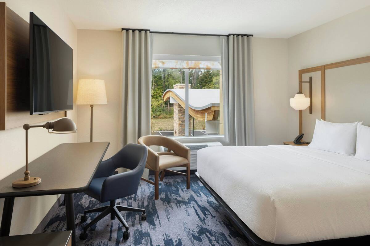 Habitación de hotel con cama, escritorio, silla, televisión, lámpara y ventana con vista a un edificio.