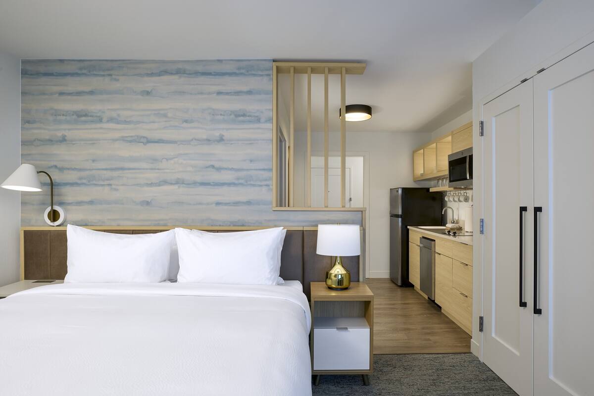 Habitación de hotel con cama, lámpara de noche, mesita de noche y vista al área de la cocina.