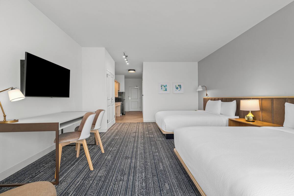 Una habitación de hotel con escritorio, dos camas, televisor y una kitchenette pequeña.