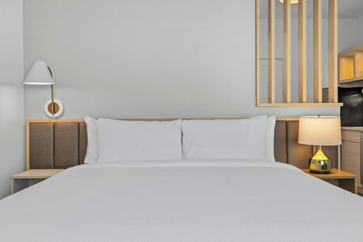 Una cama bien hecha con sábanas blancas, un cabecero de madera, una mesita de noche con una lámpara y un espejo fijado a la pared.