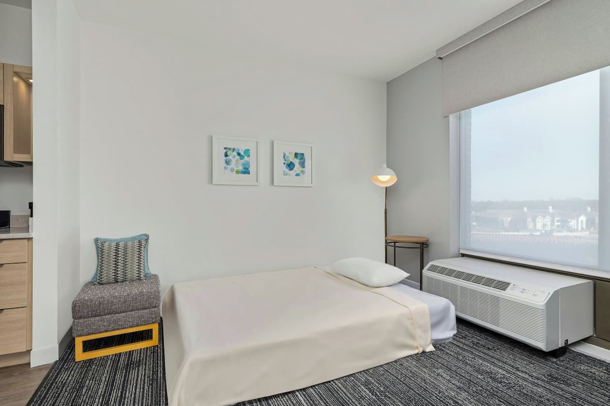 Habitación de hotel moderna con cama, un pequeño rincón de descanso, ventana con vistas a la ciudad y aire acondicionado de pared.