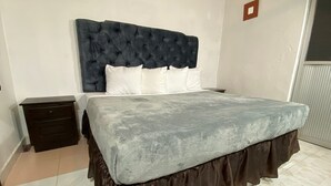 Un dormitorio con cabecera capitonada, cama con colcha gris, estructura de cama marrón con falda y mesita de noche de madera, además de suelo de baldosas.