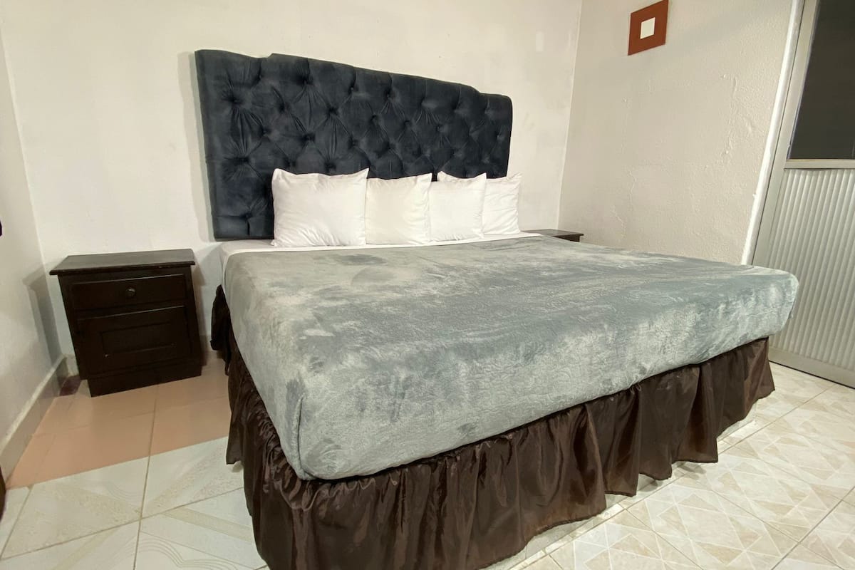 Un dormitorio con cabecera capitonada, cama con colcha gris, estructura de cama marrón con falda y mesita de noche de madera, además de suelo de baldosas.