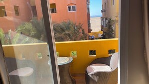 A balcony with wicker furniture and a small table, overlooking a building with balconies and palm trees. - Home from home 1 bedroom apartment with AC & WiFi in fantastic Al dora compound (Red Sea Governorate)