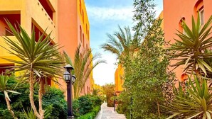 A pathway flanked by buildings with balconies, surrounded by lush greenery and a stone-tiled walkway. - Home from home 1 bedroom apartment with AC & WiFi in fantastic Al dora compound (Red Sea Governorate)