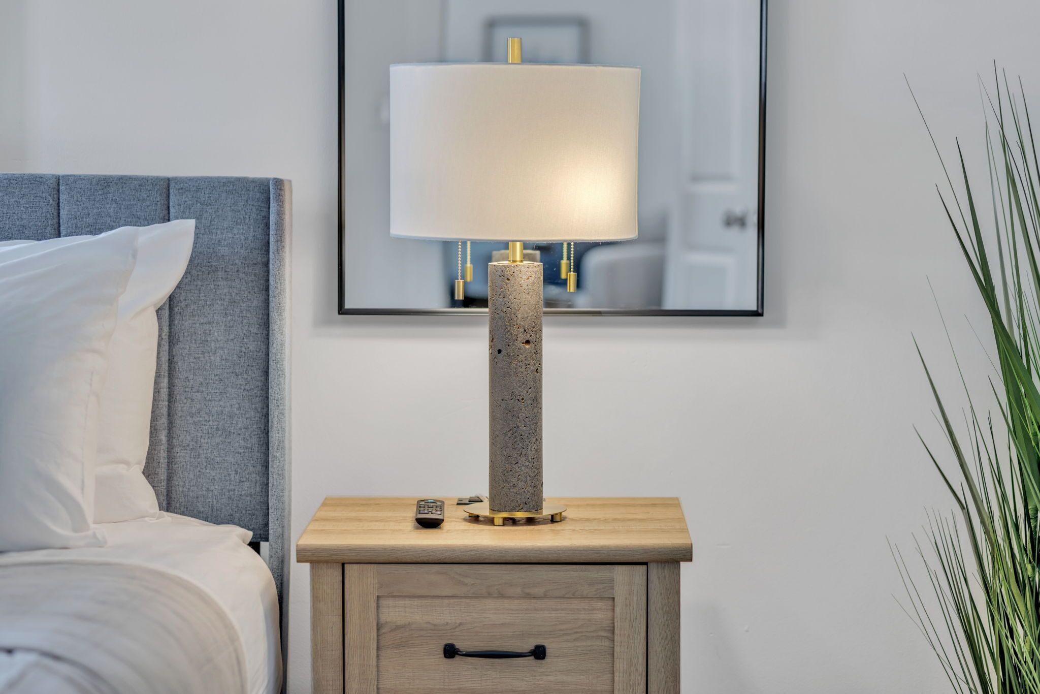 A bed with white linens, a grey headboard, and a bedside table with a lamp, remote control, and a small drawer.