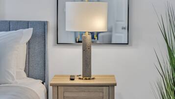 A bed with white linens, a grey headboard, and a bedside table with a lamp, remote control, and a small drawer.