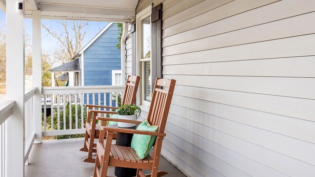 Eine Veranda mit zwei Holzstühlen und einem Tisch, einem Blumentopf und Blick auf ein blaues Haus im Hintergrund.