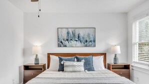 Ein Schlafzimmer mit einem großen Bett, zwei Nachttischen mit Lampen, einem an der Wand hängenden abstrakten Gemälde und einem Fenster mit Jalousien.