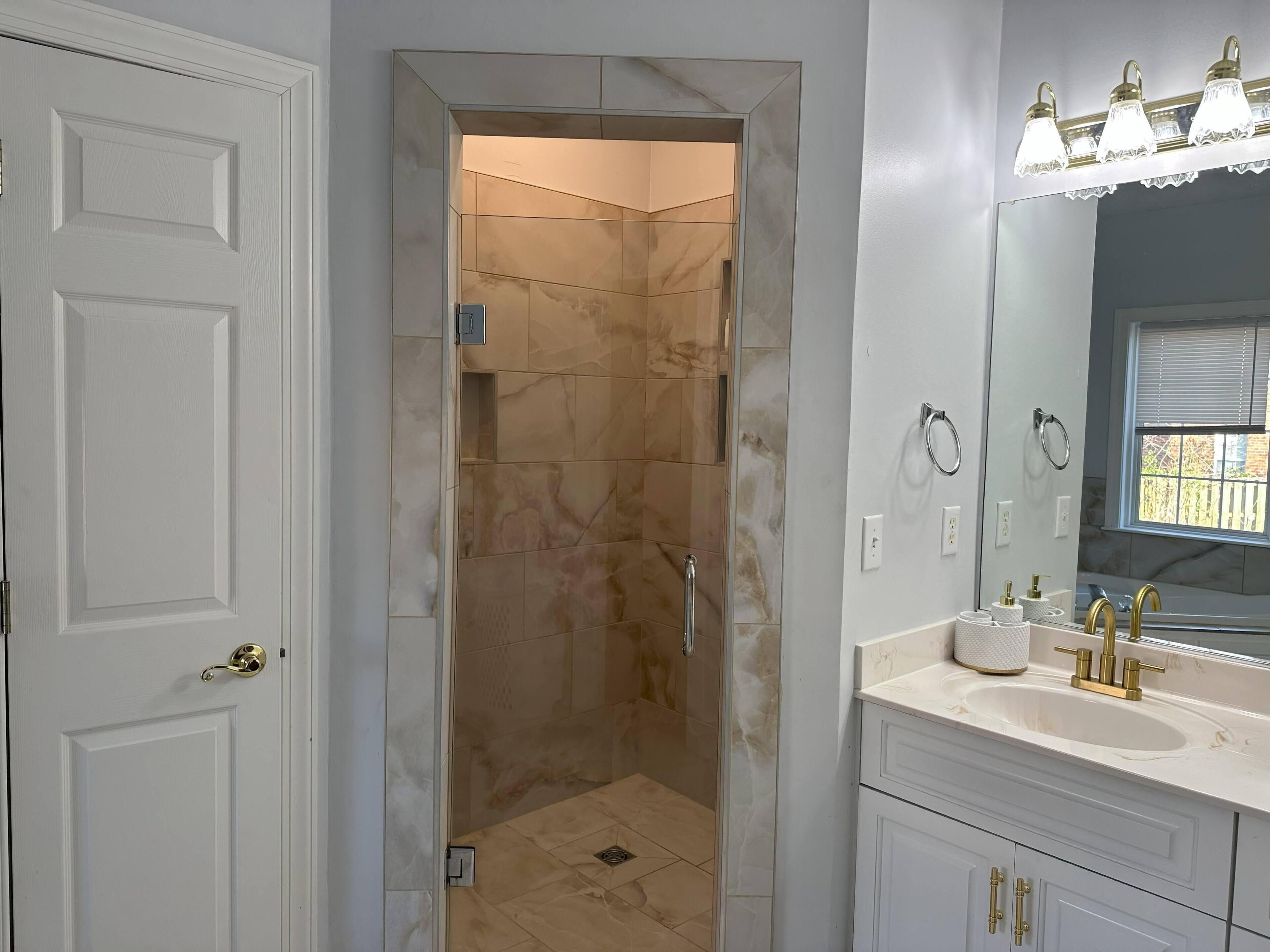 A modern bathroom with a walk-in shower, a vanity with a large mirror, and a window with blinds.