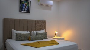 Una cama bien hecha con un edredón blanco y varias almohadas, una lámpara de noche y un cuadro en la pared.